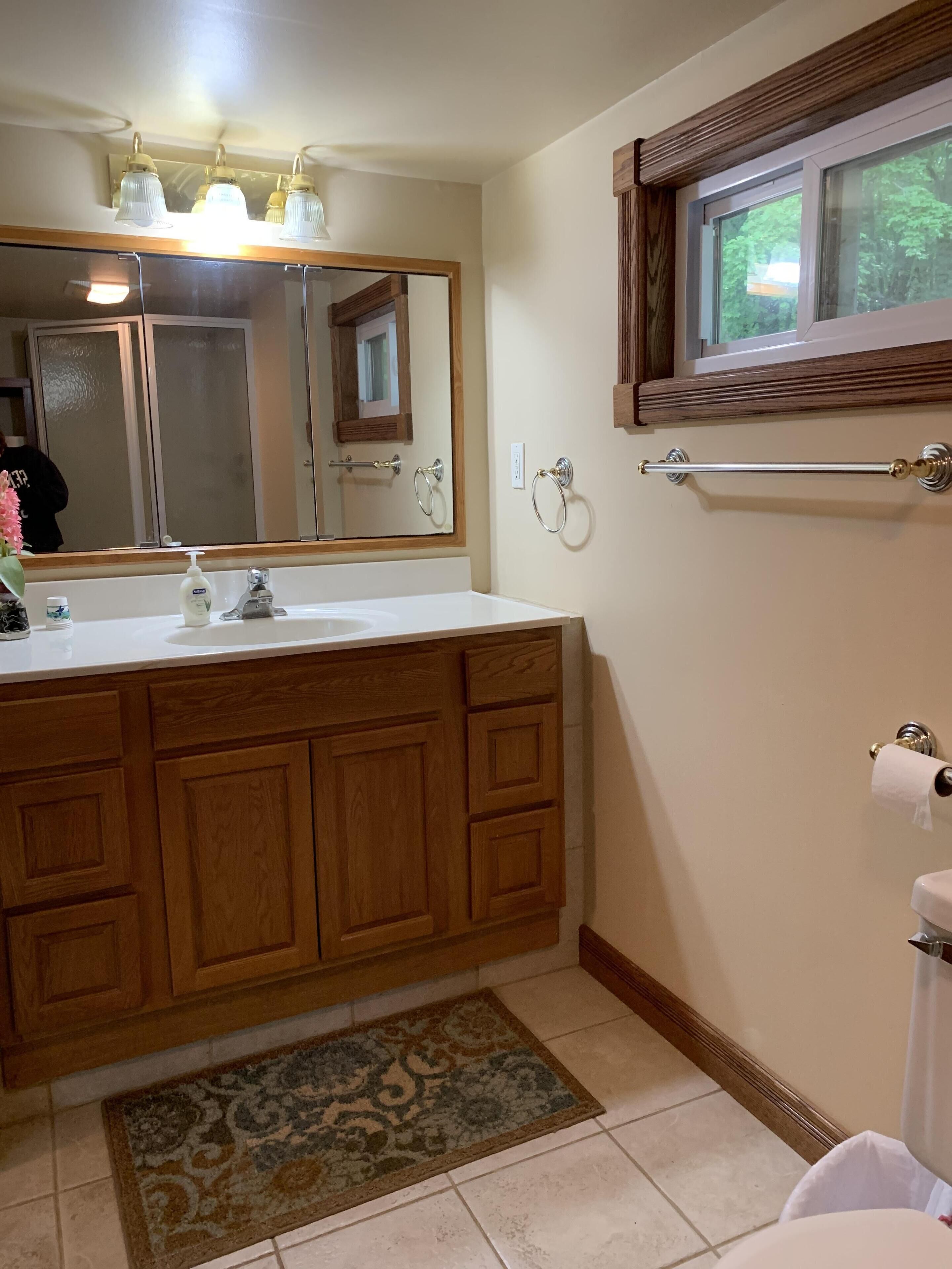 A bathroom with a wooden vanity, a large mirror, a window, and a towel rack.