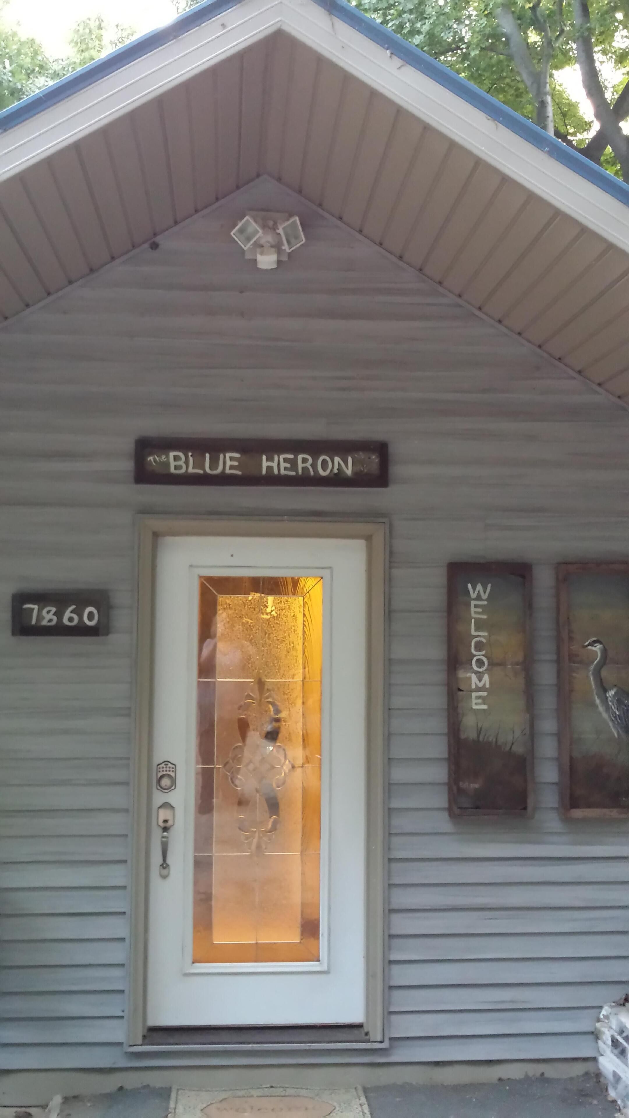 A small building with a sign reading "BLUE HERON" above the door. There are two framed signs on the wall, one with "WELCOME" and another with a swan.