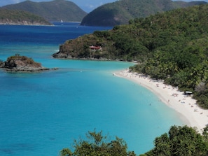 Ein Küstenstrand mit klarem türkisblauem Wasser, einem Sandufer und üppigen grünen Hügeln im Hintergrund.