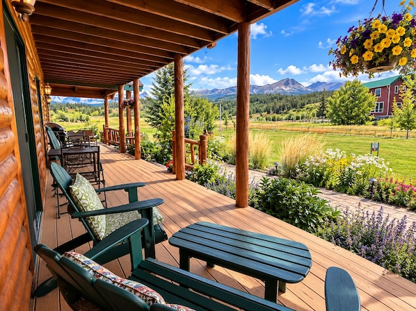 A wooden deck with outdoor seating, a hanging flower basket, and a view of a green landscape with mountains in the distance.