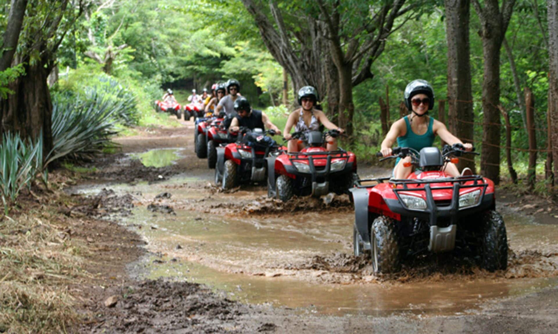 Un groupe de personnes circule en quads sur un chemin boueux à travers une forêt.