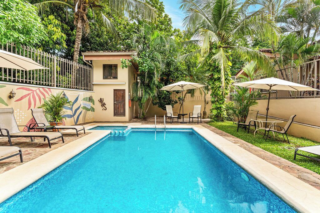 Une piscine entourée d’une végétation luxuriante et d’une clôture, avec des chaises longues et des parasols à proximité.