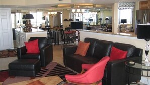 A living room with a black leather sofa, a red armchair, and a glass coffee table. There is a kitchen in the background with a dining area. - Luxurious Oceanfront Condo Ocean Views throughout in prestigious Maisons-sur-Mer (Myrtle Beach)
