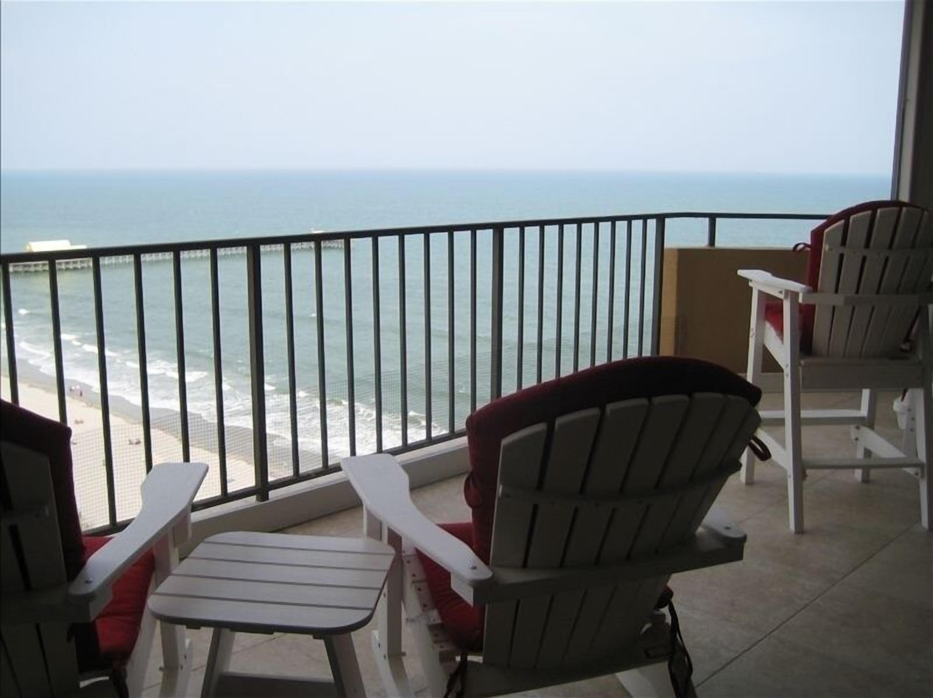 A balcony with a view of the ocean, featuring two Adirondack chairs and a small table.
