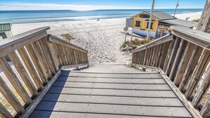 En trätrappa leder ner till ett område vid stranden, med en gul byggnad i bakgrunden.