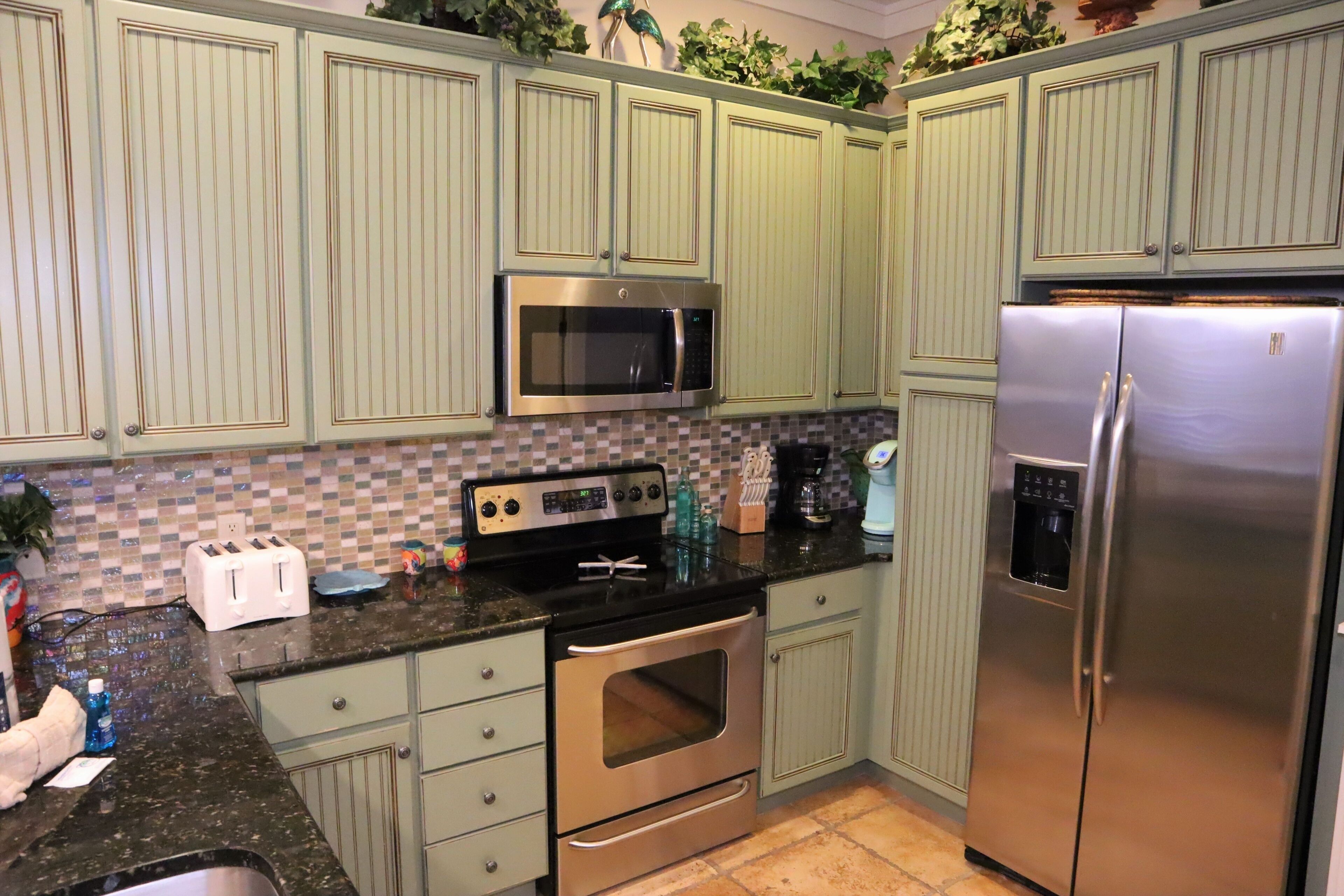 A kitchen with green cabinets, a stainless steel microwave, a toaster, and a refrigerator.