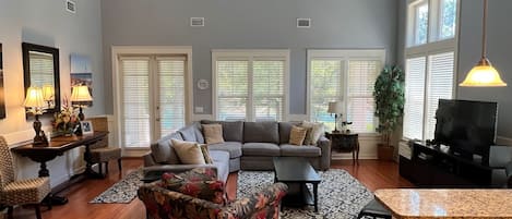 Amplia sala de estar con un sofá gris grande, un sillón floral y un televisor. Varias ventanas con persianas, piso de madera y ventilador de techo.