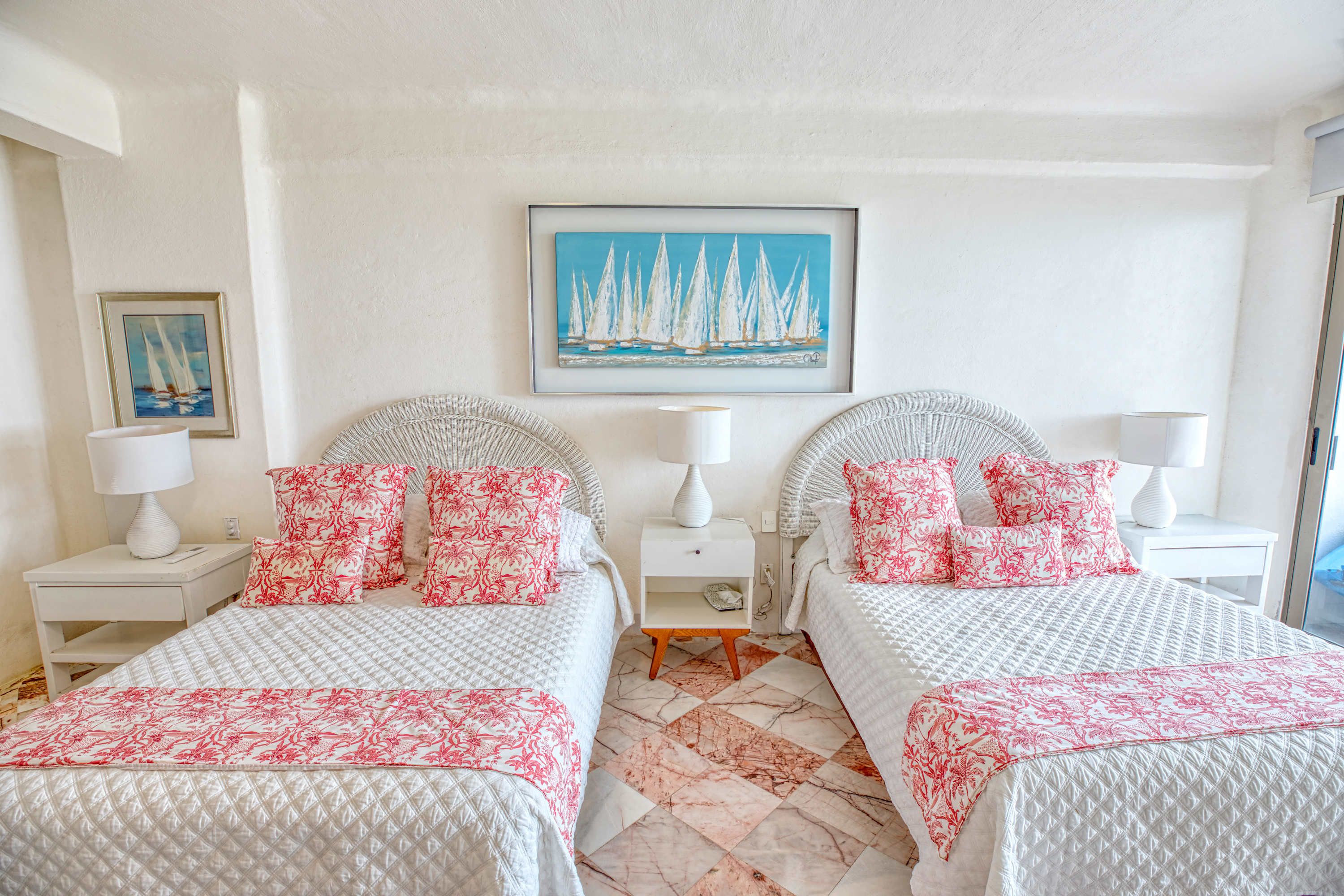 Quarto de hotel com duas camas, roupa de cama branca, almofadas vermelhas, um quadro de barcos à vela e piso de mármore.
