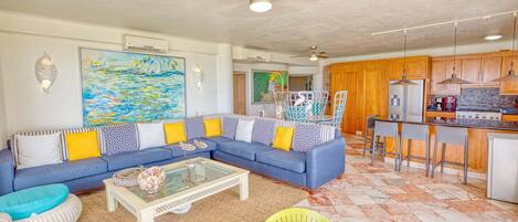 A living room with a blue sofa, a glass coffee table, and a kitchen area with wooden cabinets and a bar.