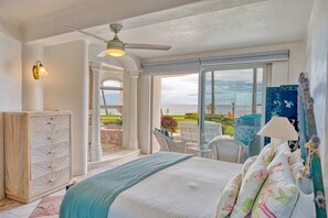 Quarto com uma cama grande, ventoinha no teto, vista para o oceano, guarda-roupa branco e varanda com móveis de exterior.