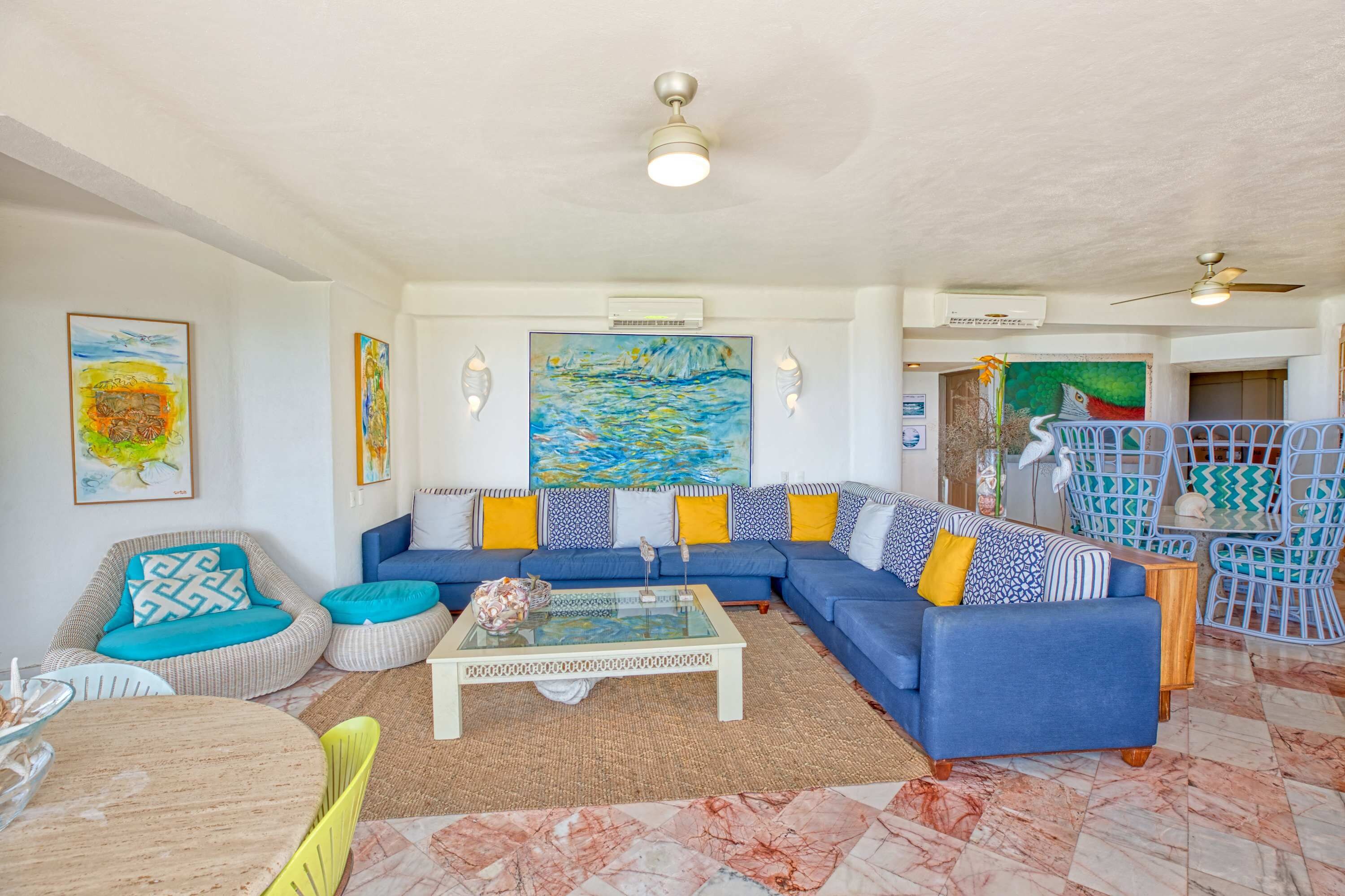 Uma sala de estar com sofá azul, almofadas coloridas, mesa de centro de vidro e um ventilador de teto.
