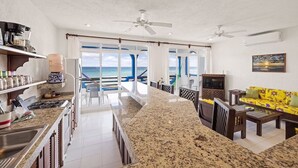 A spacious kitchen with a view of the ocean, a dining area with a yellow sofa, and a TV on a cabinet. - Absolute Beachfront - 3 Bedroom Condominium at Del Sol (Akumal)