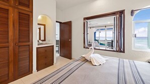 A bedroom with a bed, a wooden wardrobe, a dresser, a mirror, and a view of the sea through a window. - Absolute Beachfront - 3 Bedroom Condominium at Del Sol (Akumal)