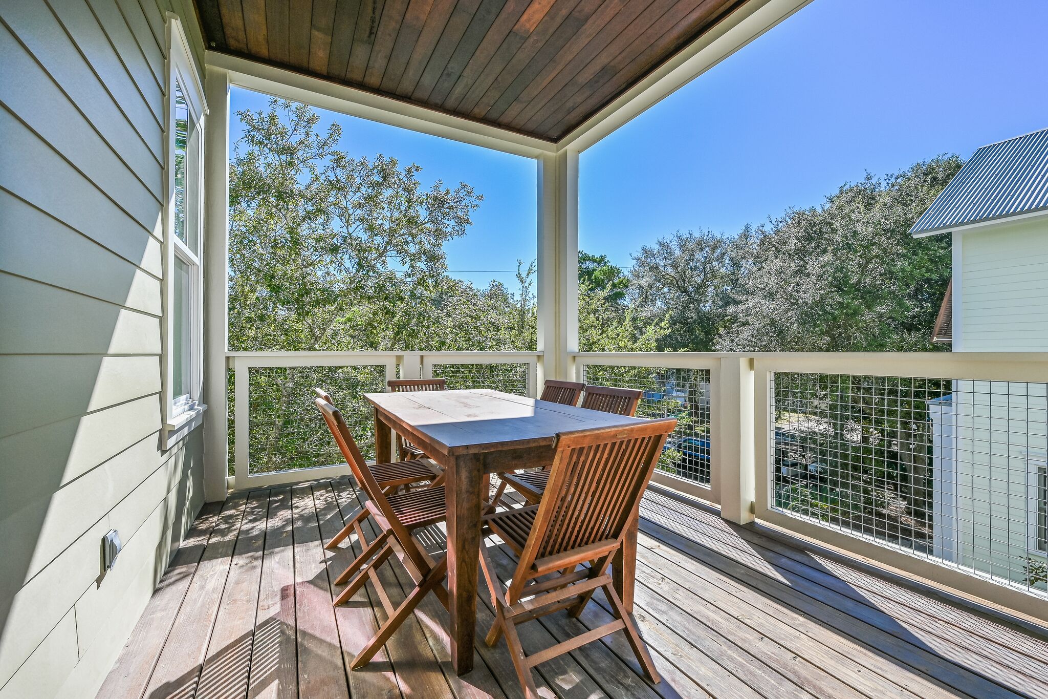 Une terrasse extérieure couverte, avec une table et des chaises en bois, entourée de rampes blanches et offrant une vue sur les arbres et une maison.