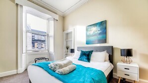 A hotel room with a large bed, a nightstand, a window with a view of buildings, and a painting on the wall. - Henderson - New Town Apartment (Edinburgh)