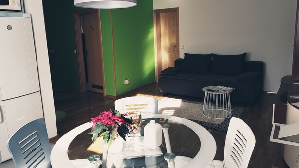 Una sala moderna con una pared de acento verde, un sofá oscuro, una mesa de comedor redonda con sillas y una mesita auxiliar con revistas.