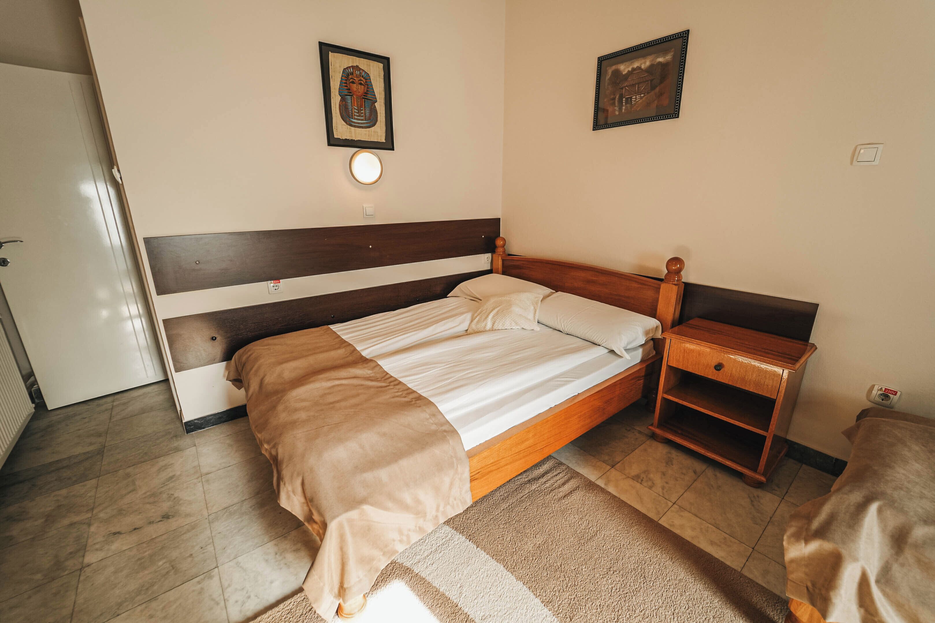 A bedroom with a wooden bed, a nightstand, a wall-mounted light, and two framed pictures.