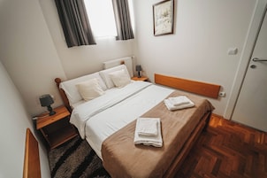 Cama bem arrumada com lençóis brancos e um espalha cama marrom, mesa de cabeceira de madeira com uma luminária, quadro na parede e chão de madeira.