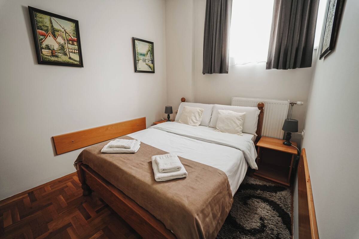 A neatly made bed with a brown bedspread and white linens, a wooden headboard, two bedside tables with lamps, and two framed pictures on the wall.
