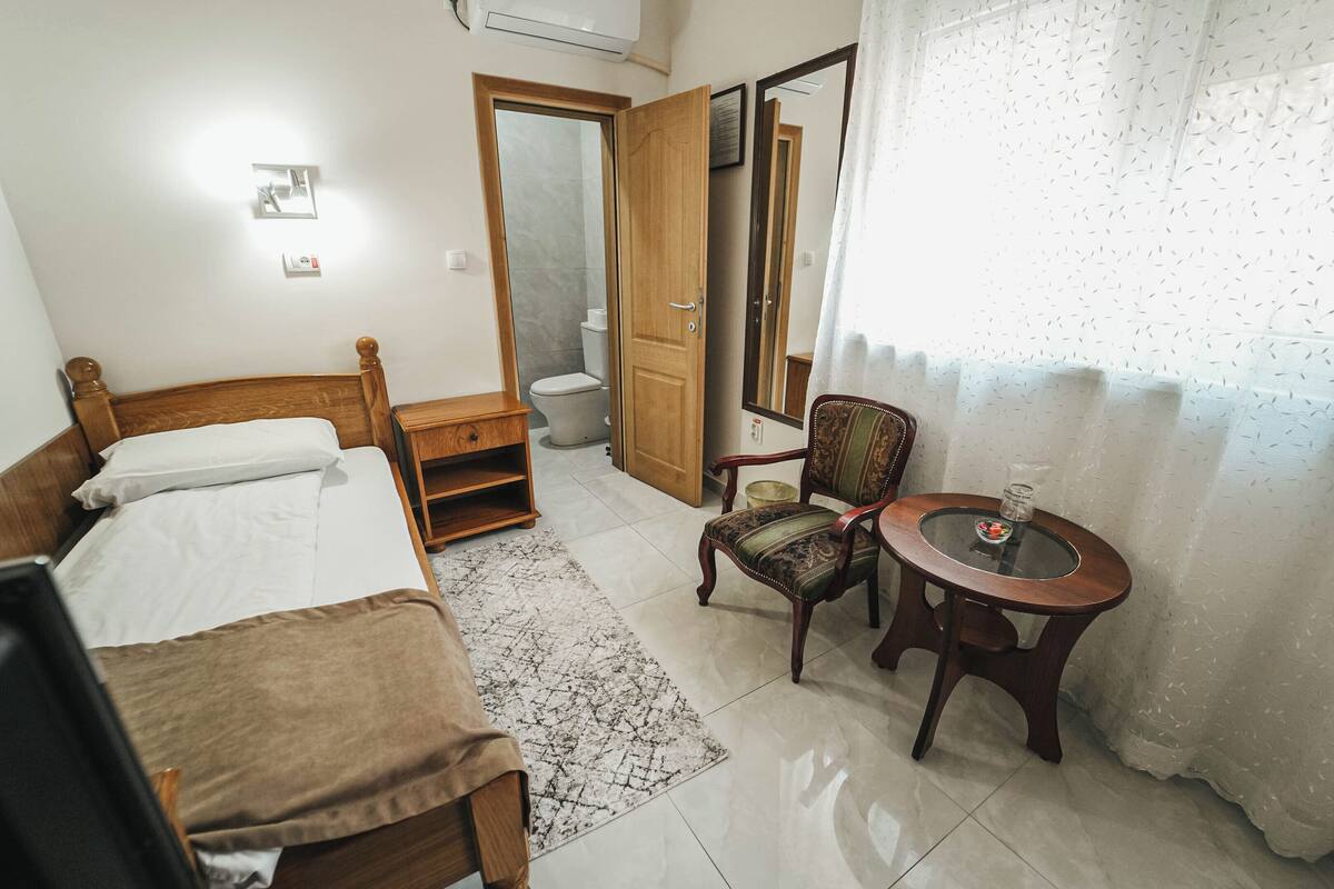A hotel room with a wooden bed, a small table with a glass, a chair, and a bathroom visible through an open door.