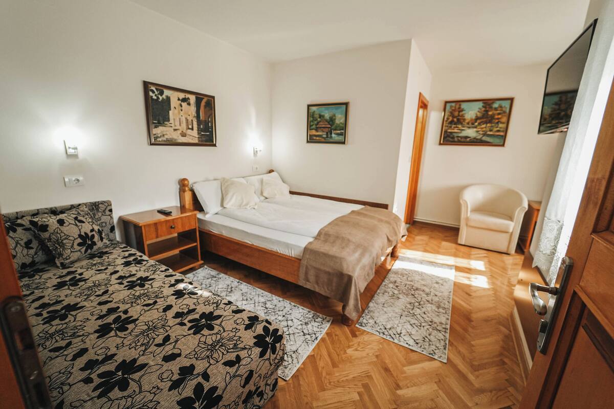 A bedroom with a bed, a sofa, a small table, and a chair. There are paintings on the walls and a TV on the right.