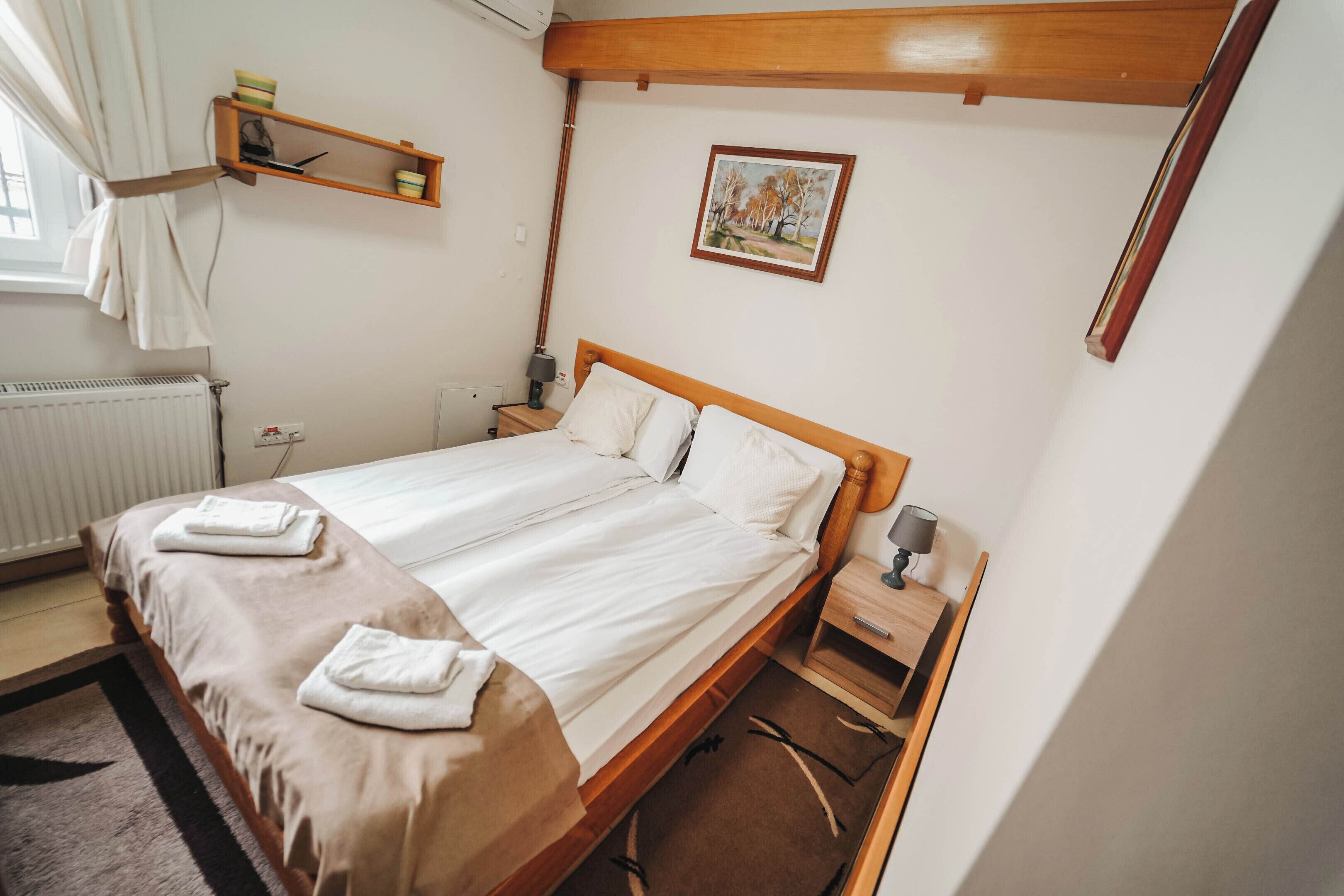 A neatly made bed with two pillows and two folded towels, a bedside table with a lamp, a wall-mounted shelf with a picture, and a radiator.