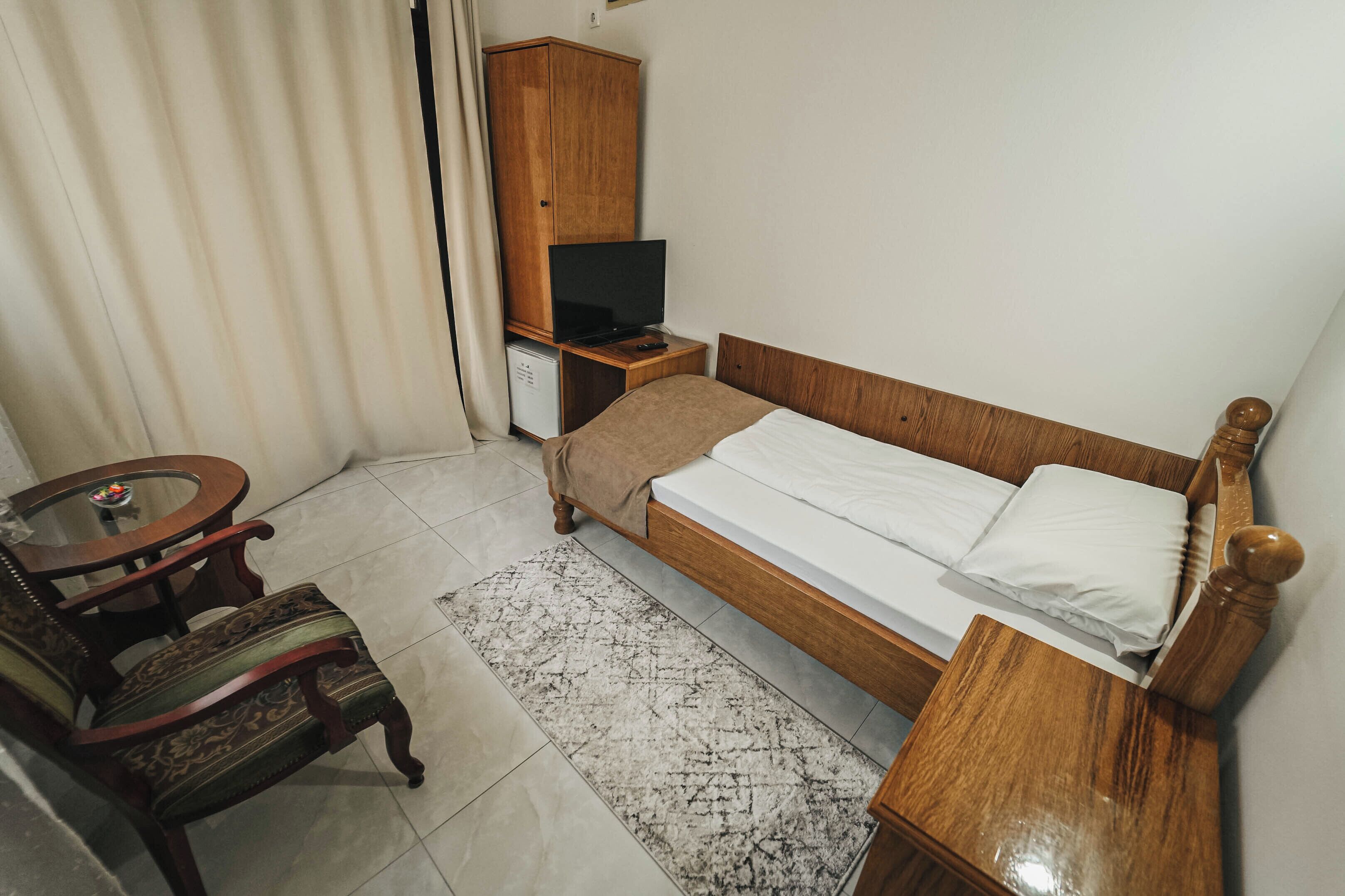 Quarto com cama de madeira, mesa de cabeceira pequena em madeira, televisão sobre um móvel, uma cadeira e uma mesinha redonda.