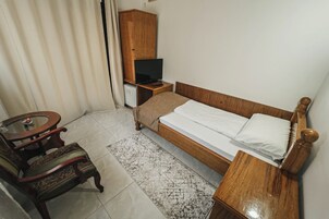 Quarto com cama de madeira, mesa de cabeceira pequena em madeira, televisão sobre um móvel, uma cadeira e uma mesinha redonda.