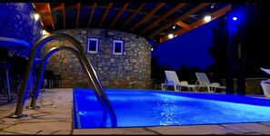 An outdoor swimming pool with a stone wall and a metal ladder. There are lounge chairs and a covered area with wooden beams.