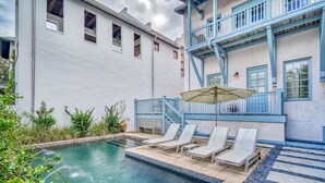 Piscina con sillas de descanso y una sombrilla, rodeada de un jardín y un edificio con barandillas azules.