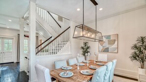 Comedor con mesa de madera dispuesta para seis personas, sillas blancas, una escalera y una lámpara colgante moderna.