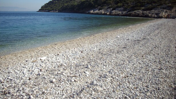 A pebble beach with clear water and a forested hillside in the background. - In the center of Ancient Greece and Saronic sea (Corinthos)
