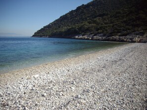 A pebble beach with clear water and a forested hillside in the background. - In the center of Ancient Greece and Saronic sea (Corinthos)
