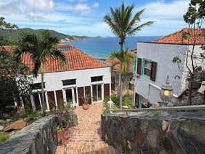 Una propiedad costera con un camino pavimentado que lleva a un edificio blanco con varias puertas, rodeado de palmeras y vistas al mar.