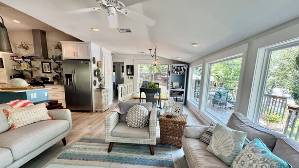 A spacious living room with a large window, a ceiling fan, and a kitchen area visible in the background. - Waterfront beauty with dock and fireplace! Check in Bay! New Hot Tub!! (Afton)