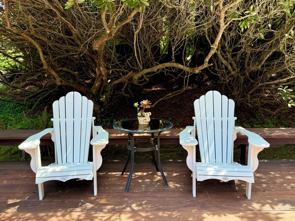 Two Adirondack chairs with a glass table and a vase of flowers between them, set on a wooden deck with a dense tree backdrop.