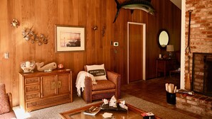 A cozy living room with a wooden wall, a large fish mounted above a door, a wicker cabinet, a chair with a beach-themed pillow, and a fireplace. - Beautiful Home in the Heart of the Majestic Redwoods.  Secluded on 10 acres. (Fort Bragg)