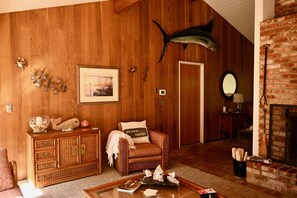 Uma sala de estar aconchegante com parede de madeira, um peixe grande montado acima de uma porta, um armário de vime, uma poltrona com almofada com tema praiano e uma lareira.