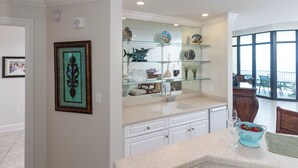 A kitchen with white cabinets, a glass display shelf with decorative items, and a view to the outside. - Beach Front, Spacious, Clean and Comfortable (Orange Beach)