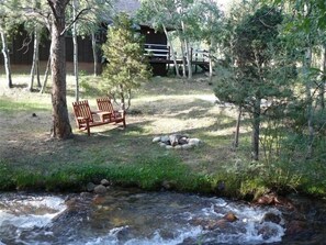 A serene outdoor area with a wooden bench, a small stream, and a rustic building in the background. - Rushing Creek w/Mountain Views! Perfect Cabin for Family/Couples Getaways! (Nathrop)