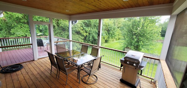 A covered deck with a glass-top table and chairs, a grill, and a view of a lush green landscape.