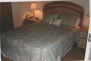 Un lit bien fait, recouvert d’une couvre-lit à motifs floraux bleus, avec une tête de lit en bois et une table de chevet sur laquelle se trouvent une lampe et un petit livre.