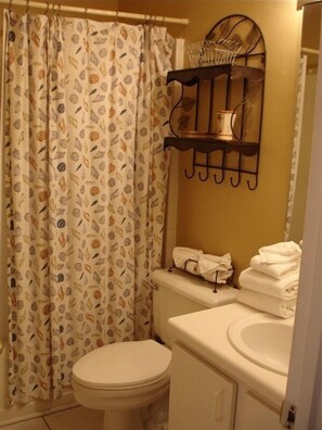 Une salle de bain comprenant des toilettes, un lavabo, un rideau de douche orné d’un motif de feuilles et un porte-serviettes avec des serviettes pliées.