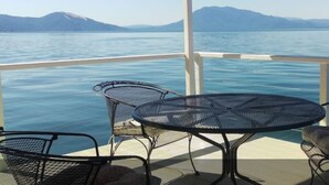 A deck with a round table and four chairs, overlooking a calm body of water with distant mountains. - Great Price! Sunset Beach Cottage with Lovely garden, beach, and boat dock! (Nice)