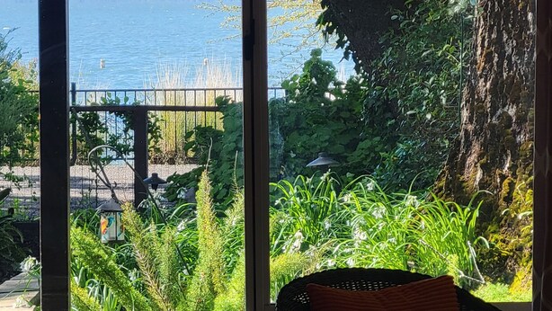 A room with a large window overlooking a body of water, a chair, and a view of a bridge. - Great Price! Sunset Beach Cottage with Lovely garden, beach, and boat dock! (Nice)