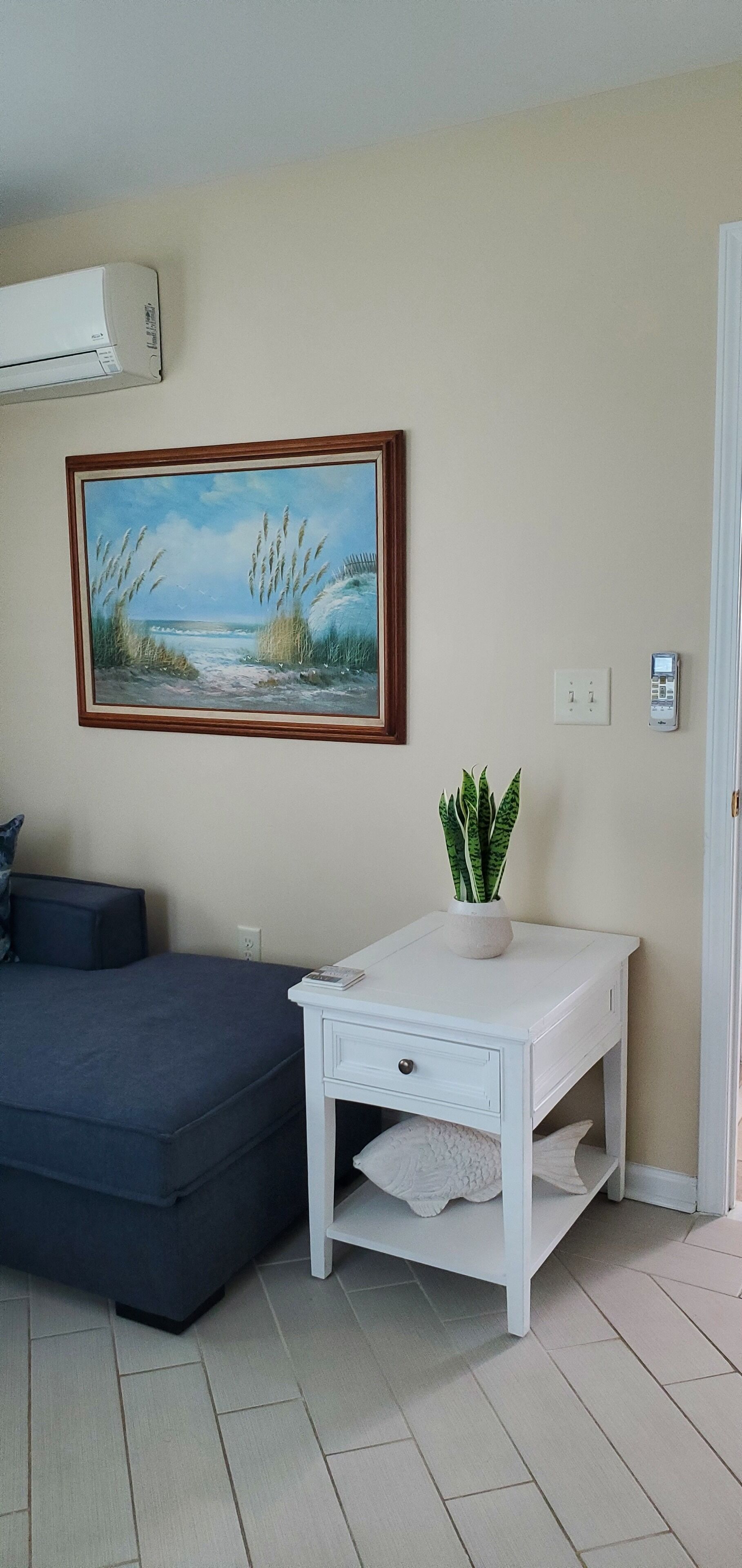 Een kamer met een donkerblauwe bank, een witte tafel met een plant, een schilderij van een strand en een airconditioner aan de muur.