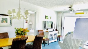 A dining area with a wooden table, wicker chairs, a white wicker chair, and a television on a white cabinet. - The Sea Palm, A Coastal Cottage Steps From The Beach!!! (Sunset Beach)
