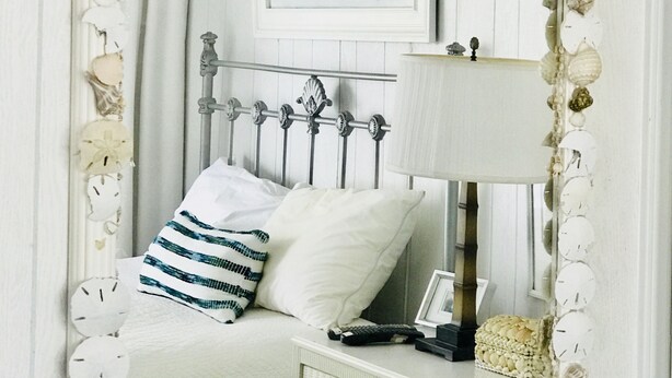 A bedroom with a bed, a nightstand, a lamp, and a mirror framed with seashells and starfish. - The Sea Palm, A Coastal Cottage Steps From The Beach!!! (Sunset Beach)