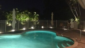 Une piscine illuminée la nuit, entourée de palmiers et agrémentée de parasols blancs.