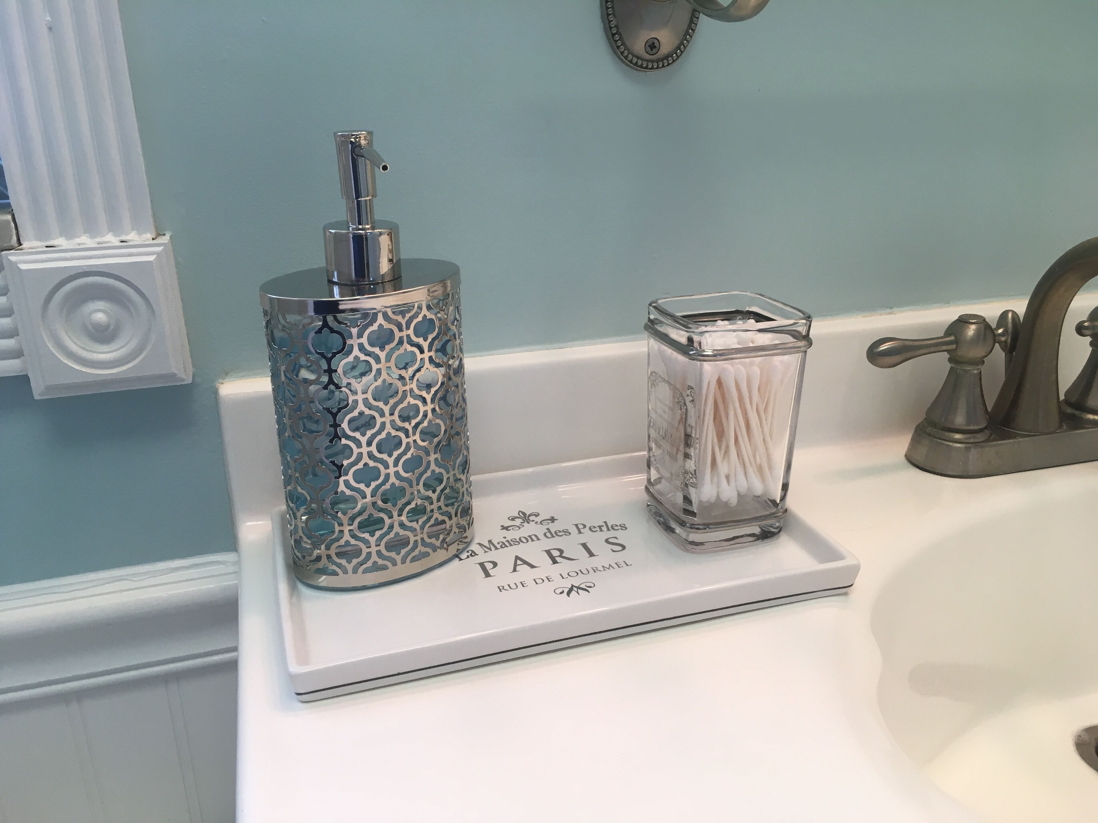 Un lavabo con encimera blanca, una botella azul y plateada, y un recipiente de vidrio con bastoncillos de algodón.